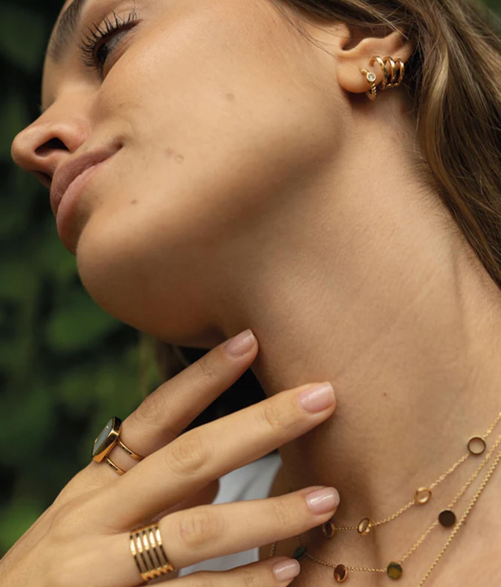 junge frau mit langen haaren neigt ihren Blick weg von der Kamera und trägt dabei goldene Halsketten, Ringe und Ohrringe von der Marke Zag Bijoux